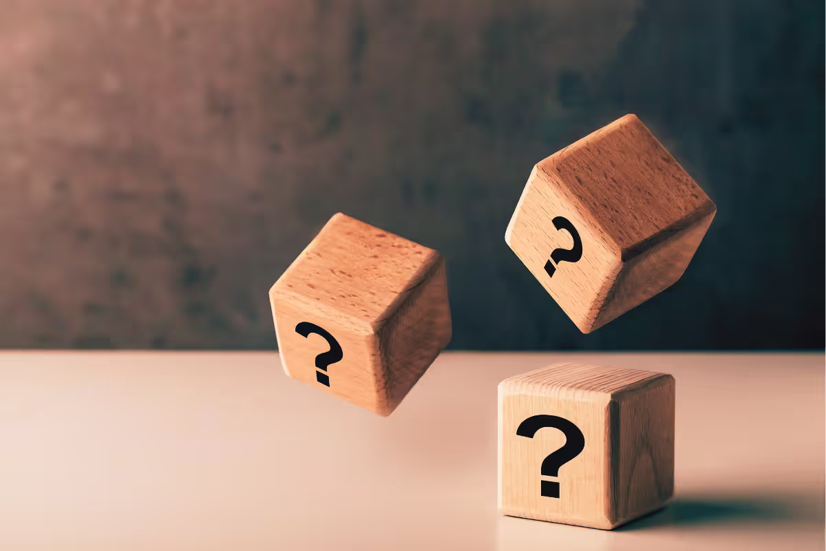 A photo of 3 wooden dice, each with a question mark on one side. One has landed on a desk and the other two are still in the air.